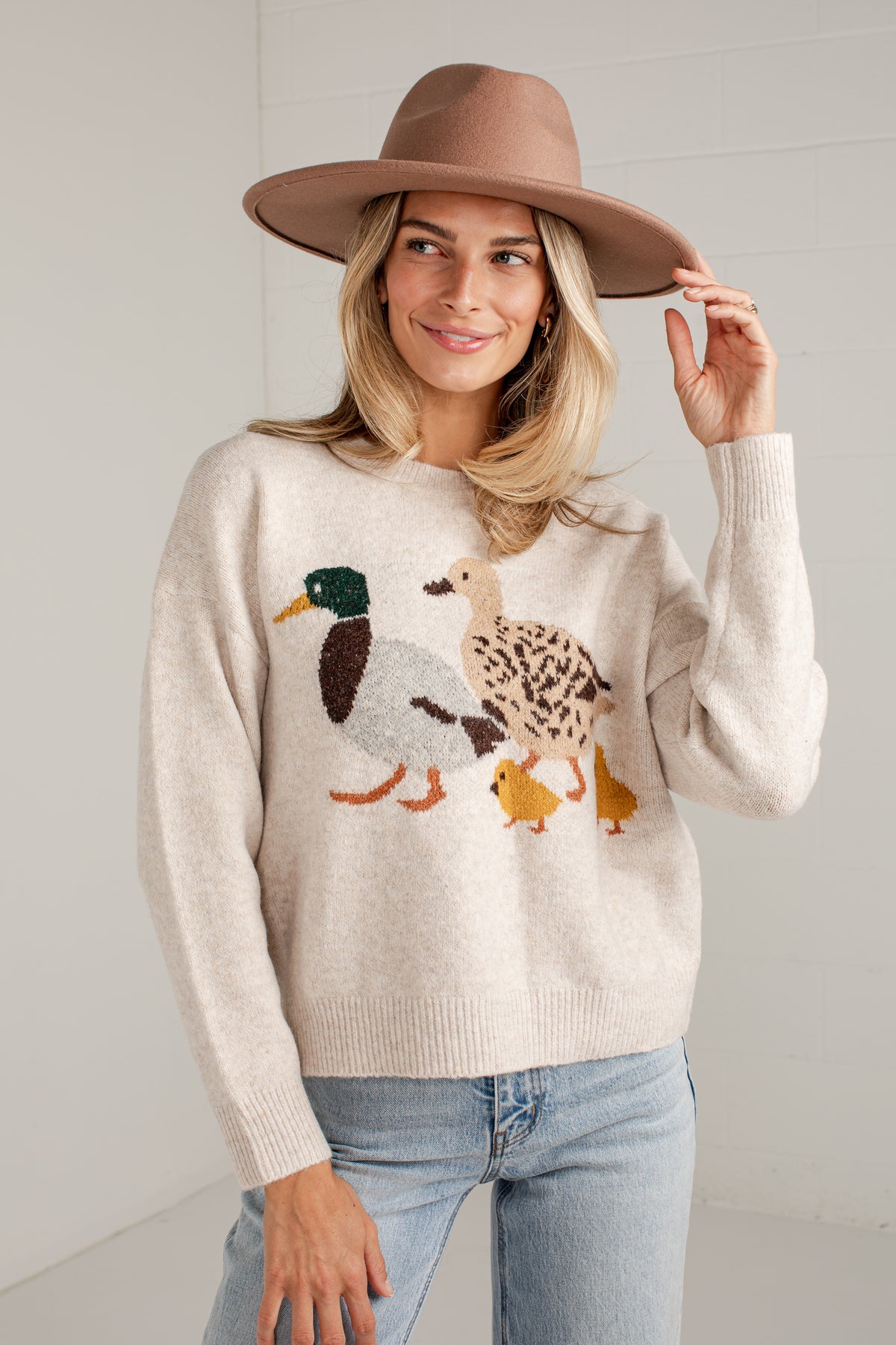 A woman wearing a cream knit sweater with a duck family motif, paired with light blue jeans and a brown wide-brim hat, standing against a white paneled wall.