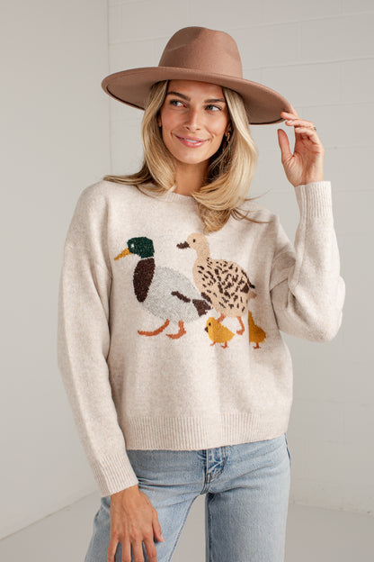 A woman wearing a cream knit sweater with a duck family motif, paired with light blue jeans and a brown wide-brim hat, standing against a white paneled wall.