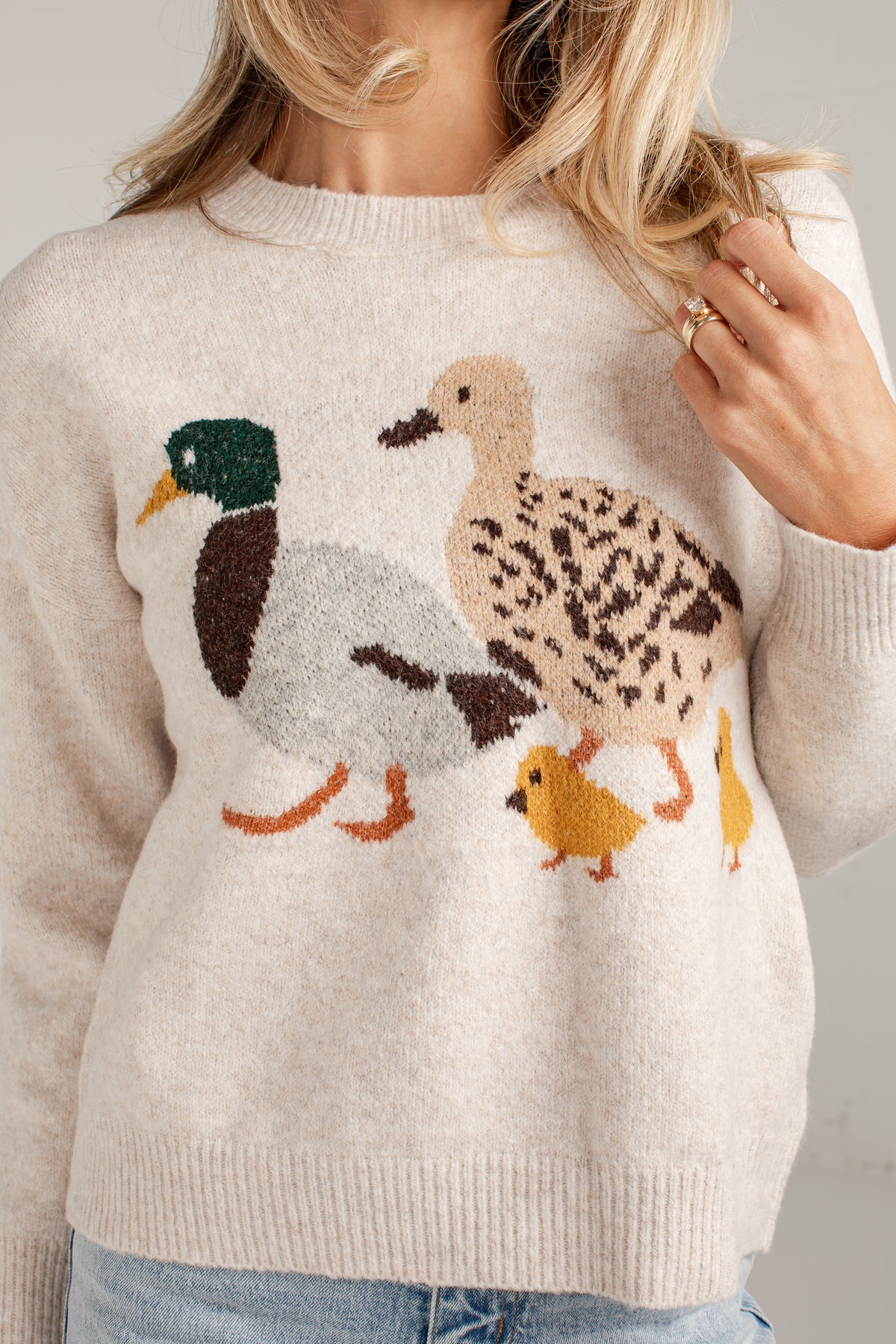 A woman wearing a cream-colored knit sweater with a detailed knitted duck family motif on the front, featuring a mallard, a speckled duck, and two yellow chicks.