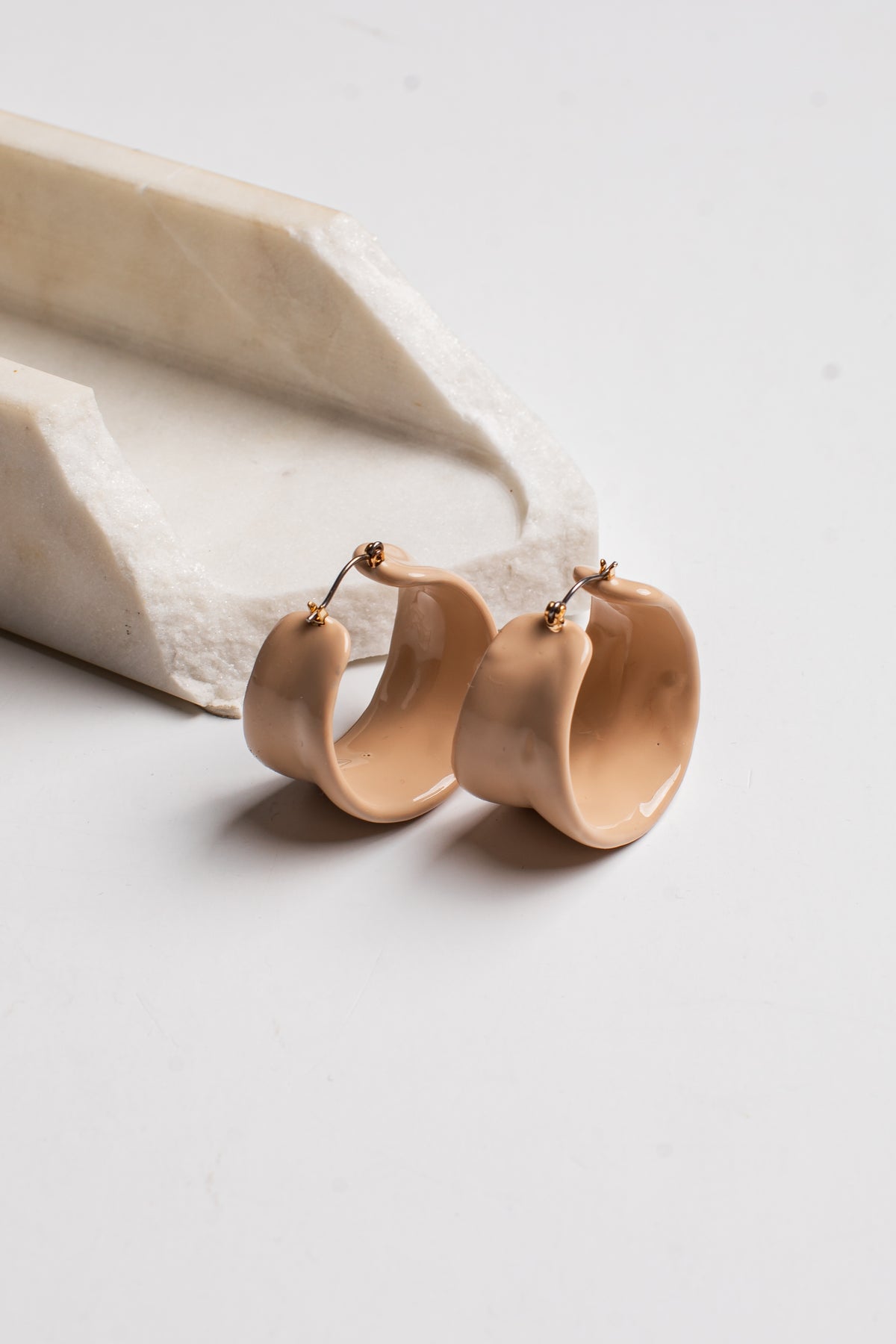 A pair of chunky, wavy hoop earrings in a glossy light beige finish with small gold post backings, displayed on a white surface next to a white stone prop.