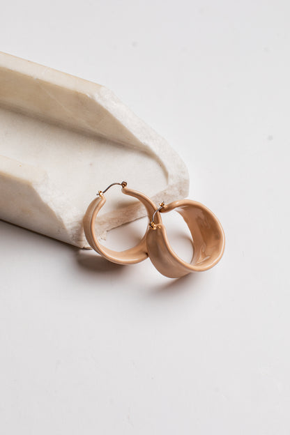 A pair of chunky, matte-finish hoop earrings in a warm beige/tan color, resting on a white stone tray against a clean white background.