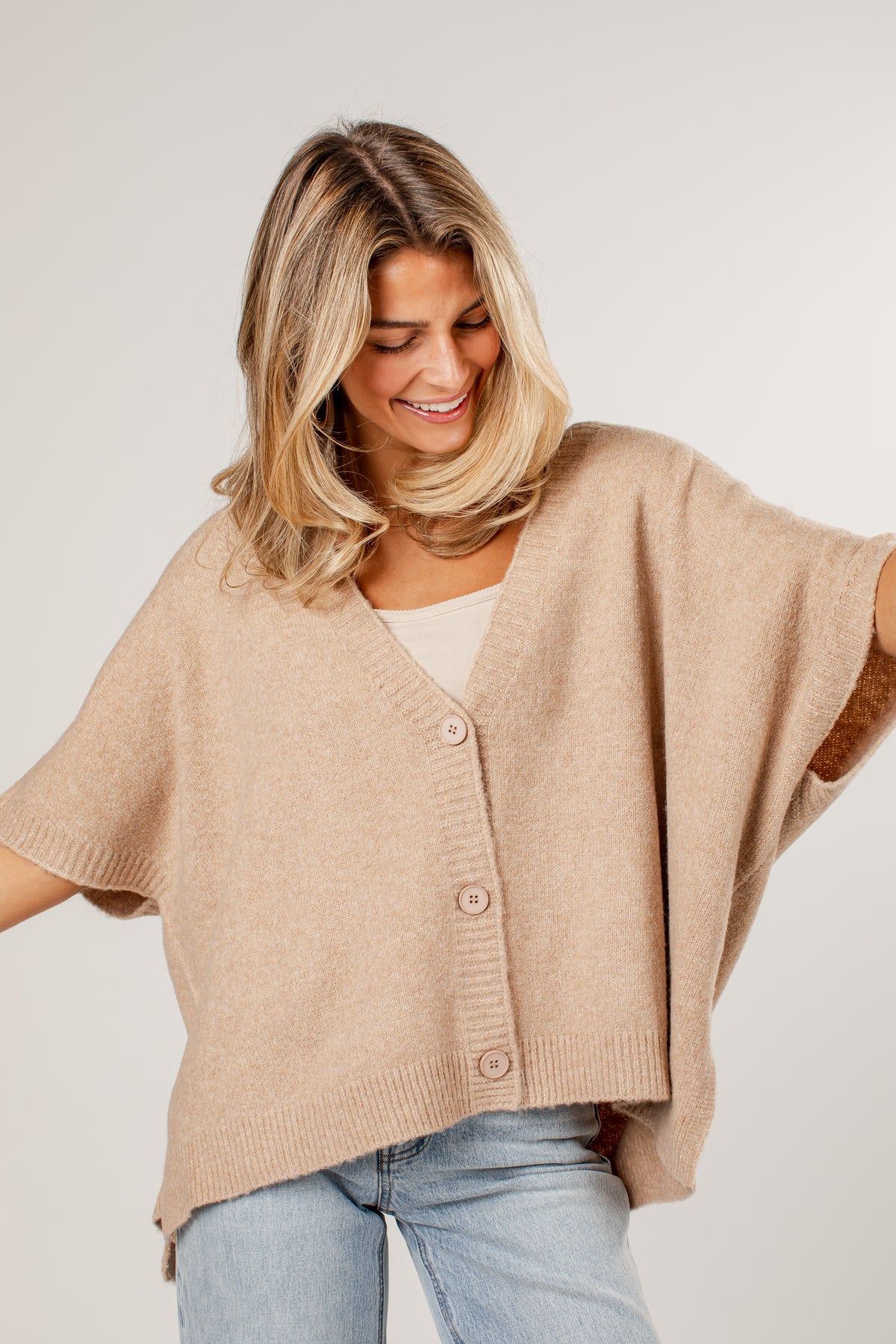 A woman wearing a beige knit cardigan with short wide sleeves and a button-front closure, paired with light blue jeans, photographed against a plain light gray studio background.