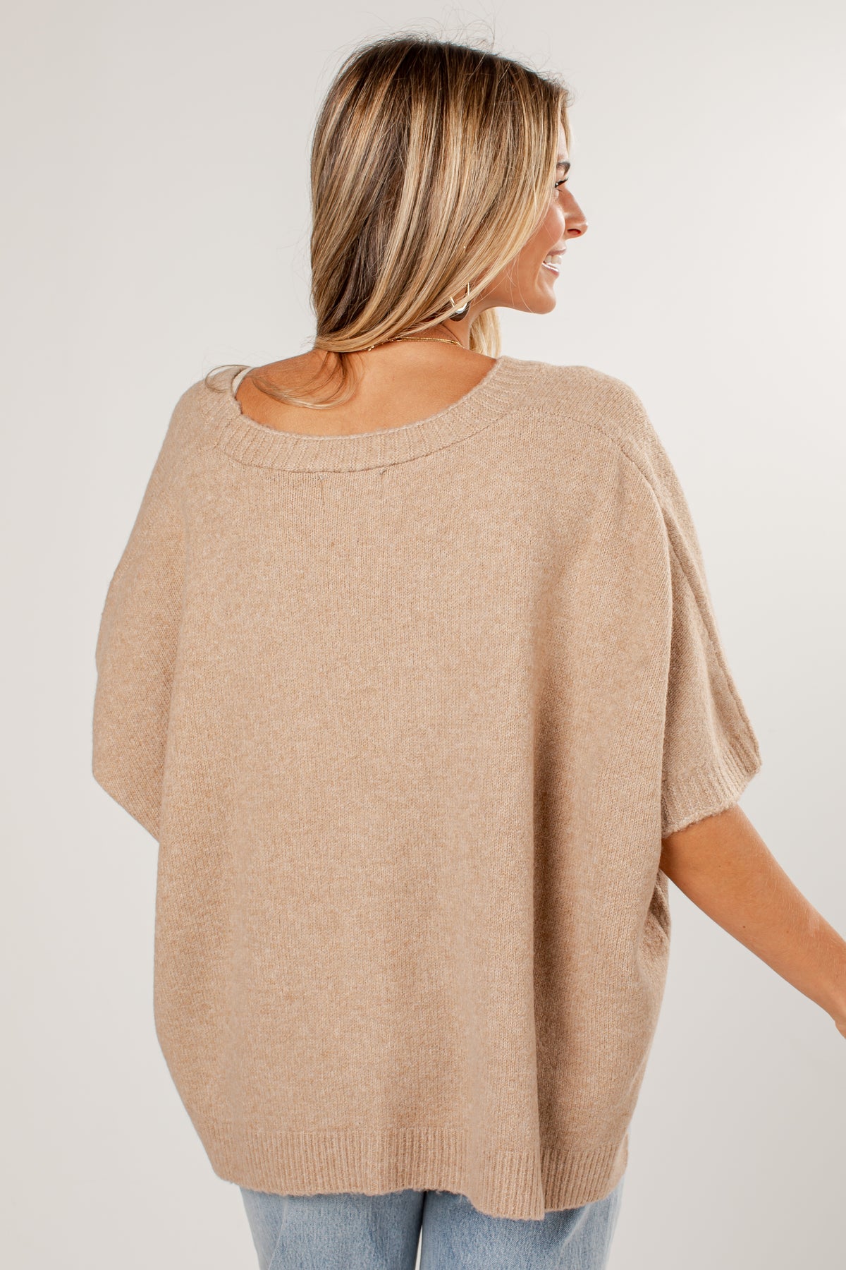 A woman shown from the back wearing a loose, short-sleeve knit sweater in a light beige or oatmeal color, paired with light blue jeans, against a plain light gray studio background.