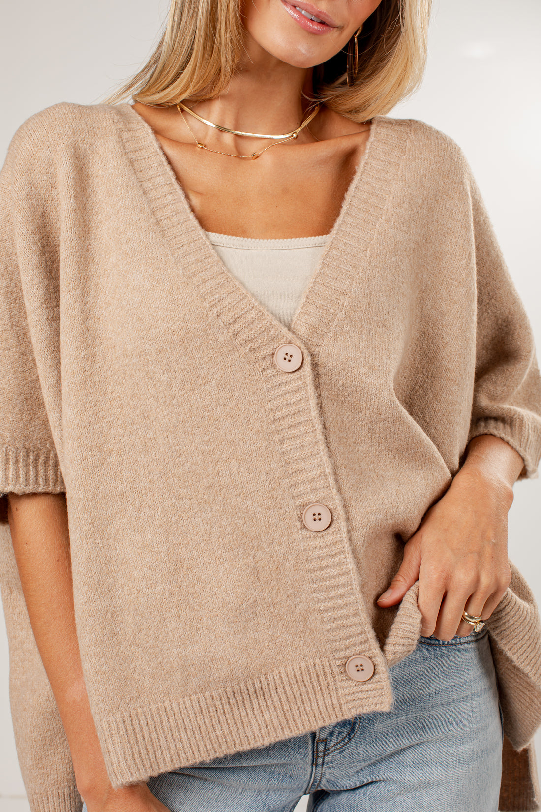 A woman wearing a beige knit cardigan with a V-neck, three-quarter sleeves, and button-front closure over a white top and light blue jeans.