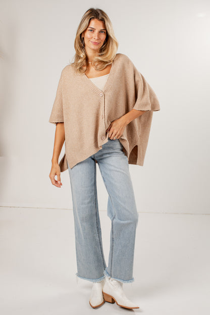 A woman modeling a light beige knit poncho-style cardigan with a button front, worn over a white top and paired with light blue jeans and white ankle boots, against a plain white studio background.