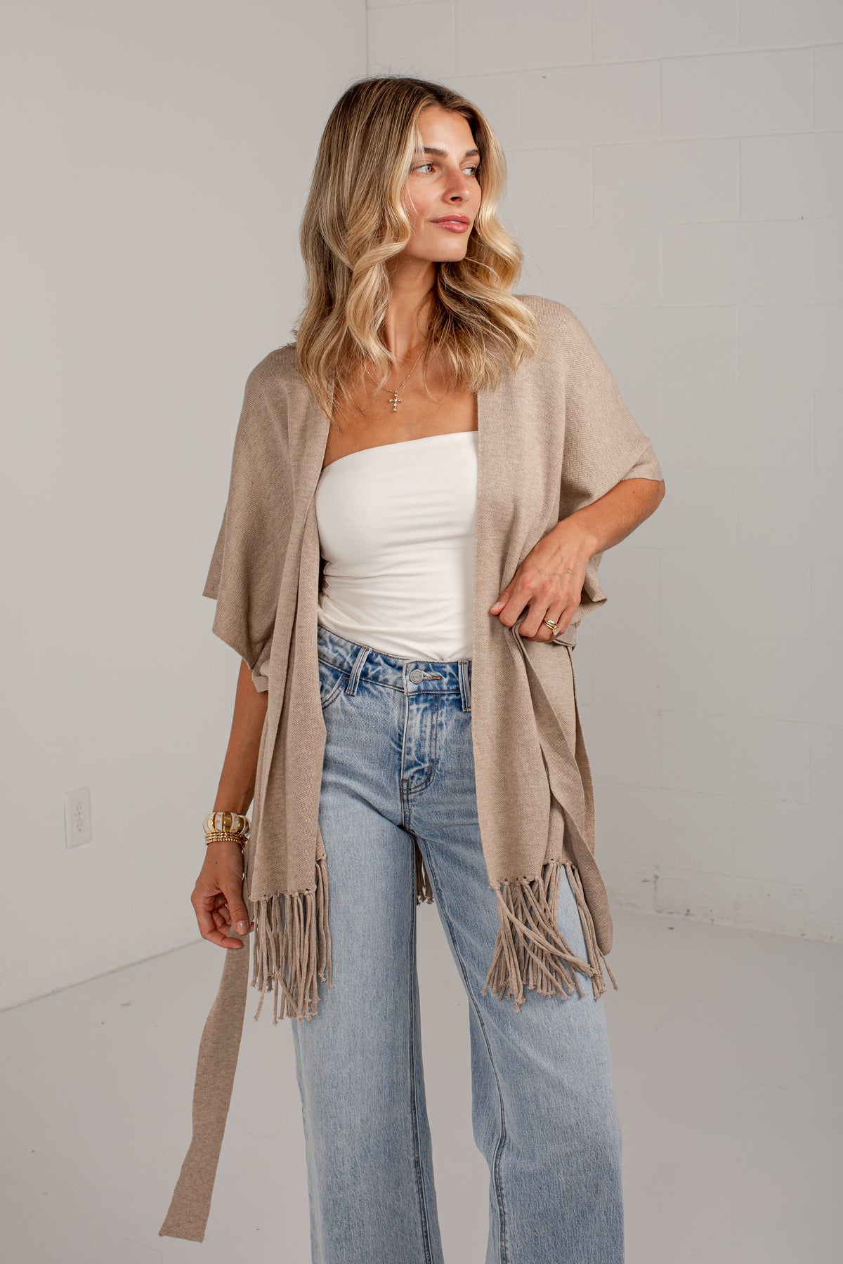 A woman models a light beige open-front fringed cardigan over a white tube top and light-wash blue jeans, standing against a white wall.