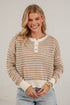 A smiling blonde woman wearing a cream and tan knit henley sweater with light blue jeans, standing against a white brick wall.