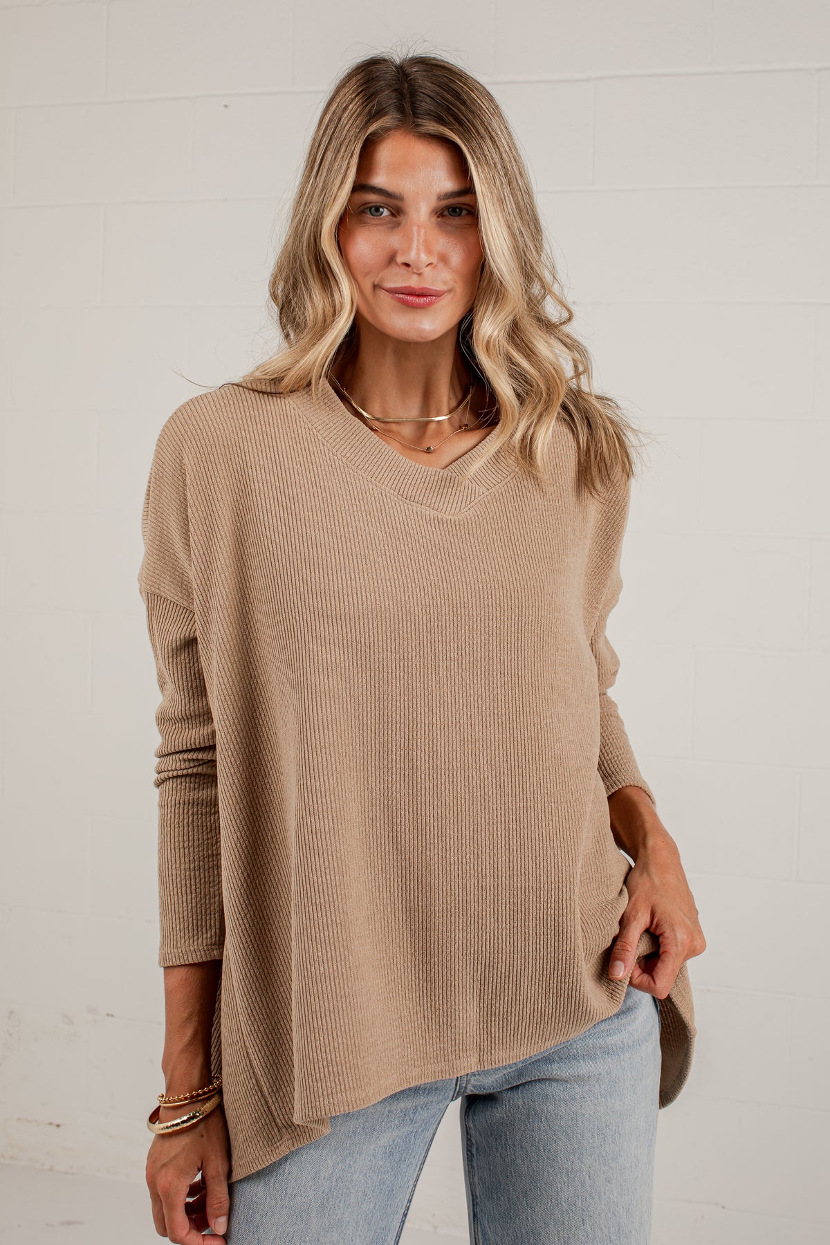 A woman wearing a beige ribbed knit long-sleeve sweater with a V-neck and relaxed fit, paired with light blue jeans, standing against a white brick wall.