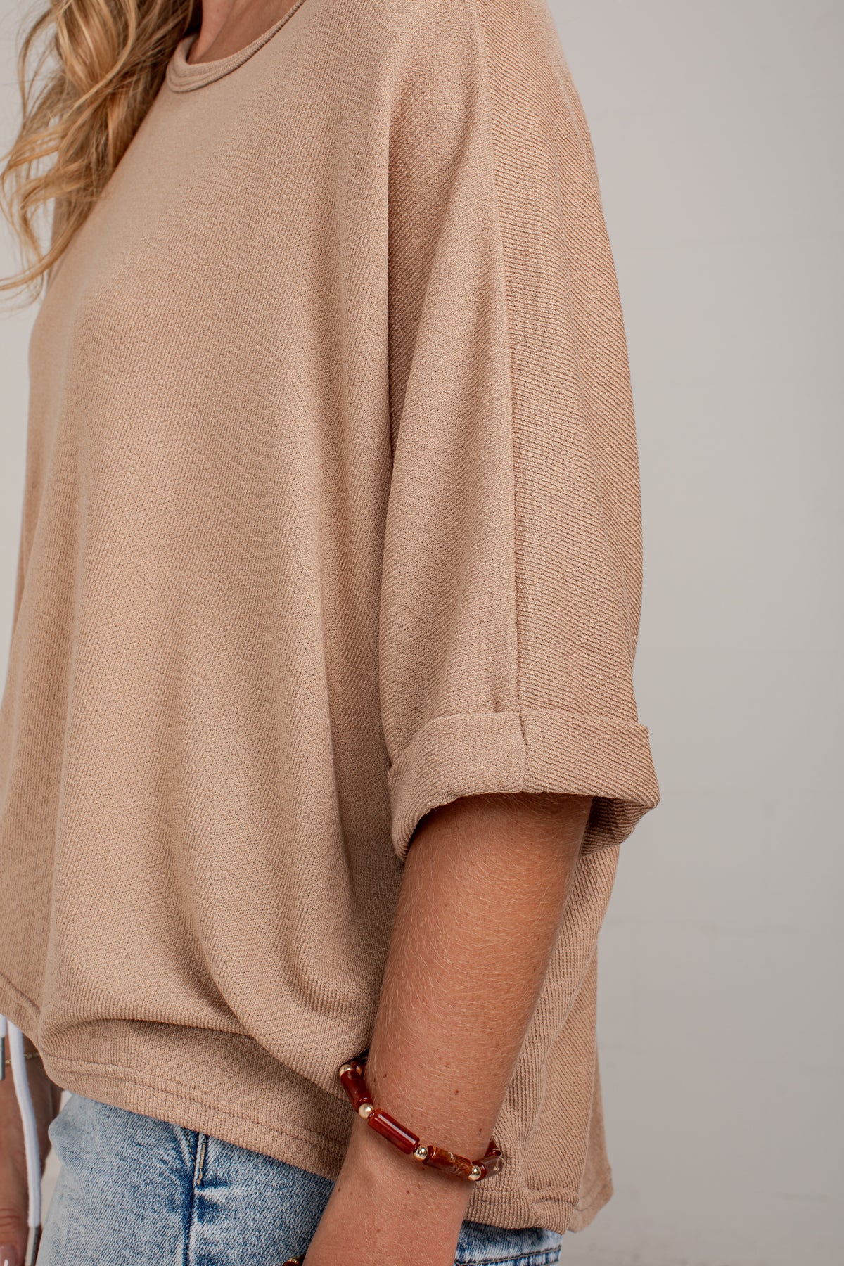 A close-up of a person wearing a loose beige knit top with rolled three-quarter sleeves and light blue jeans against a plain light gray background.