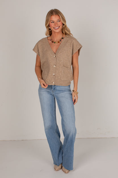 A woman modeling a beige short-sleeve button-front knit top with front pockets, paired with light-wash blue jeans, standing against a plain light gray studio background.
