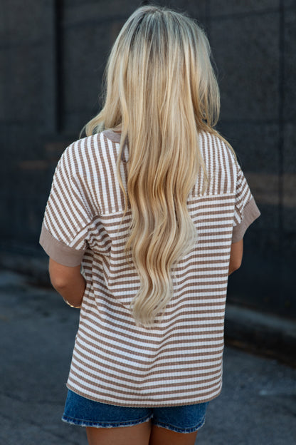 A woman viewed from behind wearing a beige and white horizontally striped short-sleeve t-shirt and denim shorts, standing outdoors against a dark gray wall.