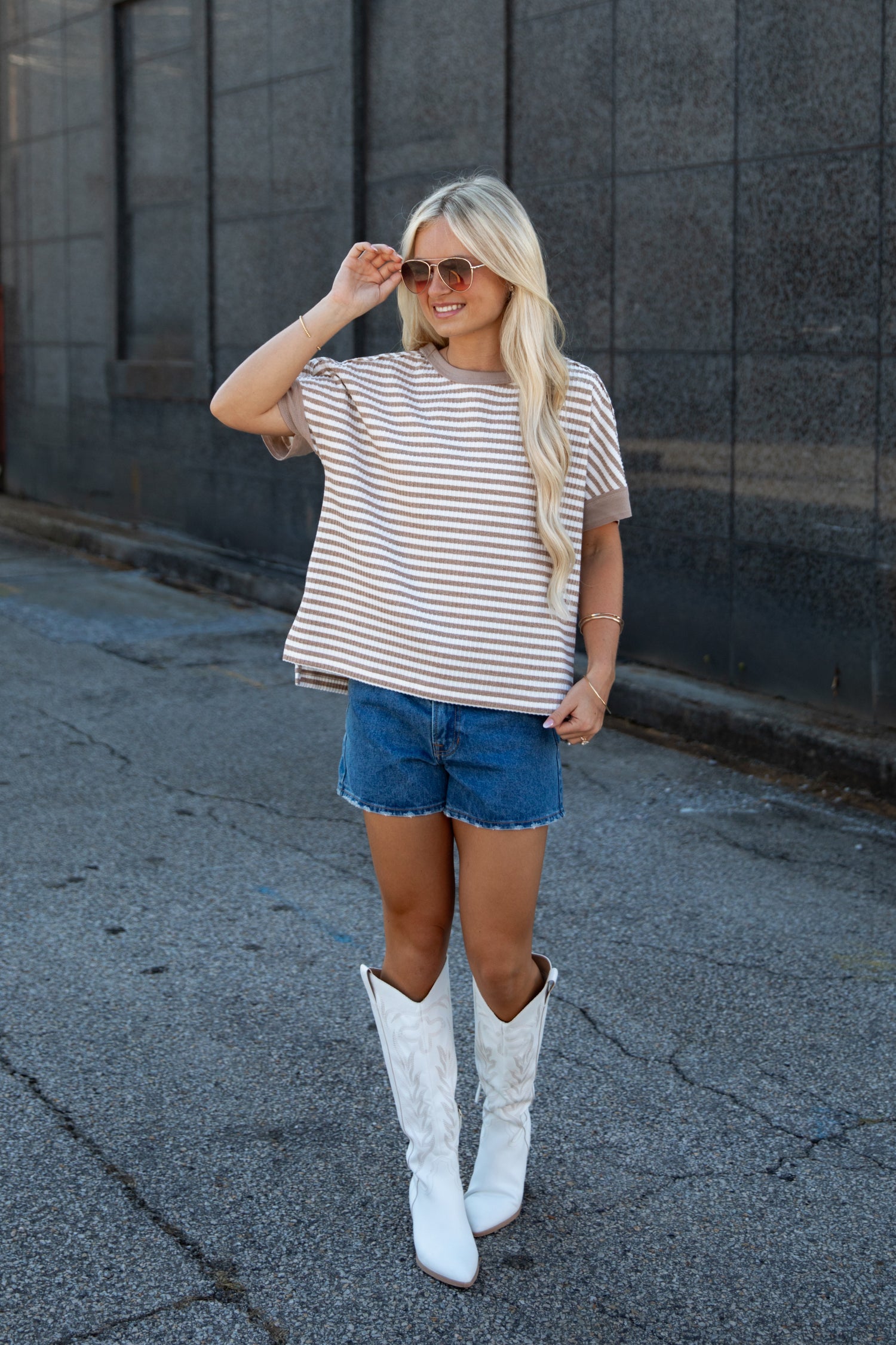 A woman stands outdoors wearing a beige and white horizontally striped short-sleeve t-shirt, blue denim shorts, and tall white cowboy boots, posing in front of a dark gray stone wall.