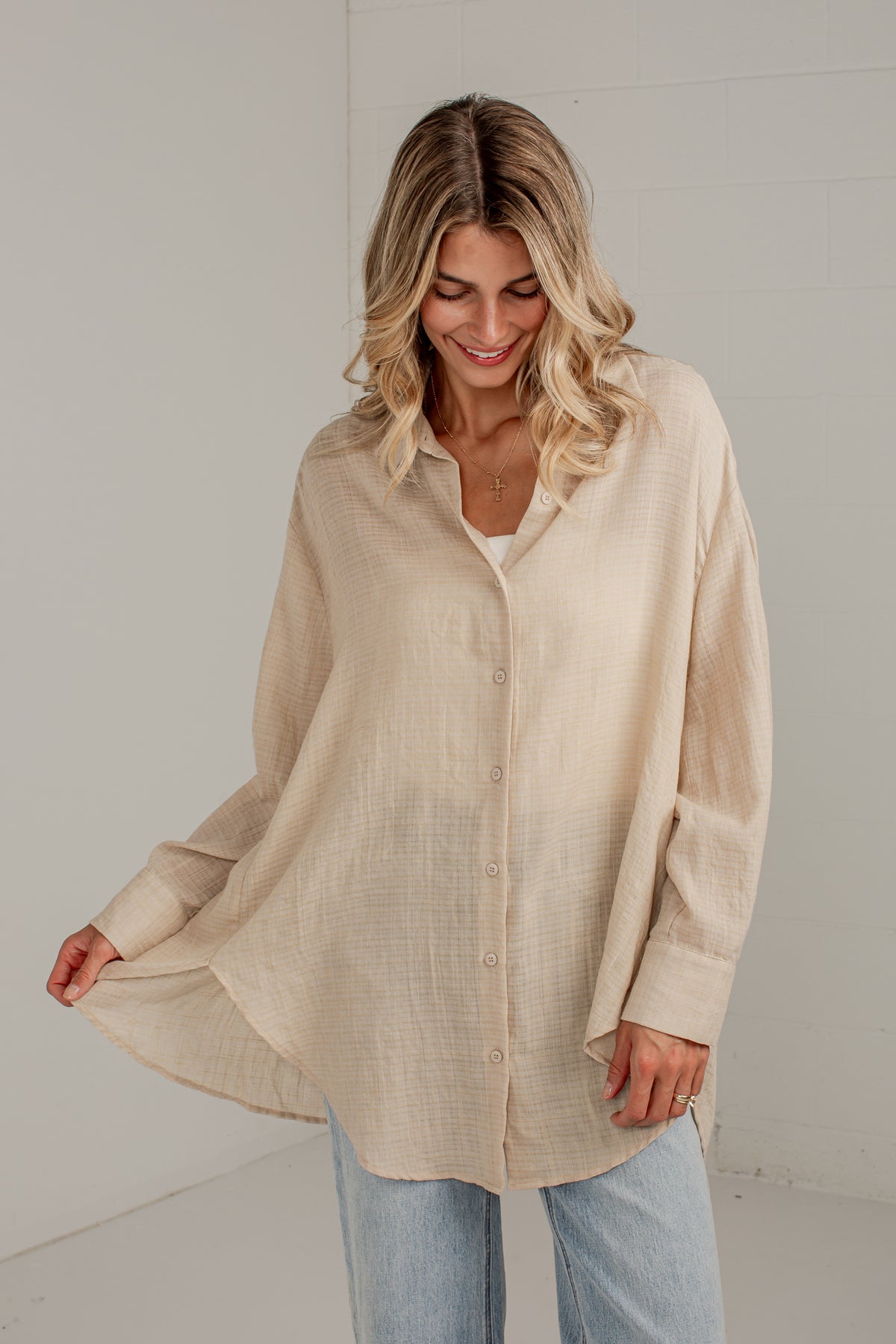 A woman wearing a light beige, oversized long-sleeve button-up shirt with a textured linen-like fabric, paired with light blue jeans, standing against a white wall.