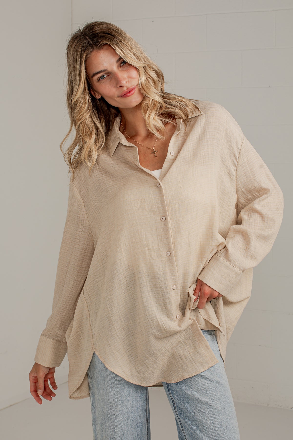 A woman wearing an oversized, light beige textured linen button-down shirt with light blue jeans, standing against a white brick wall.