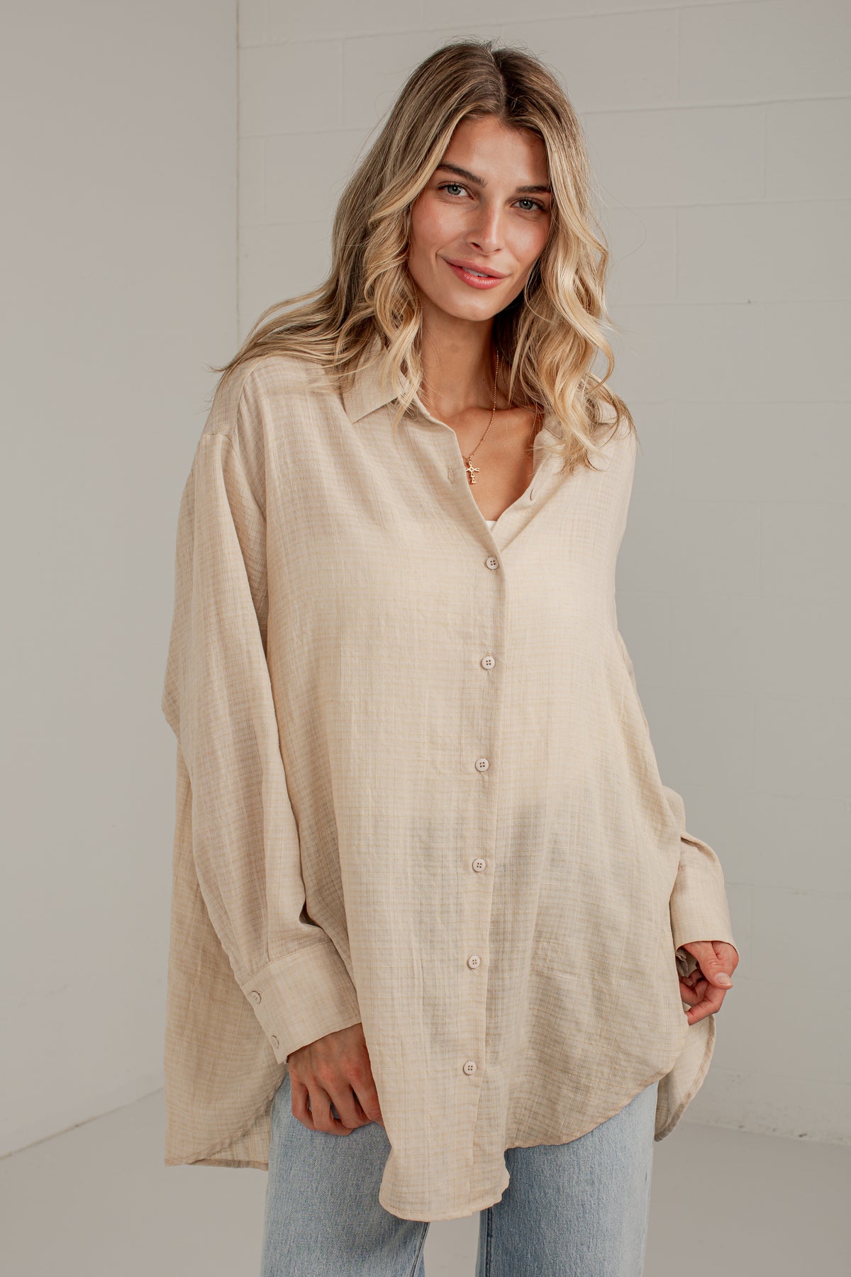 A woman wearing a light beige, oversized long-sleeve linen button-up shirt and light blue jeans, standing against a neutral light gray wall.