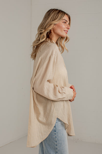 A woman wearing a loose, light beige linen long-sleeve top with a high-low hem and light blue jeans, standing against a white brick wall.