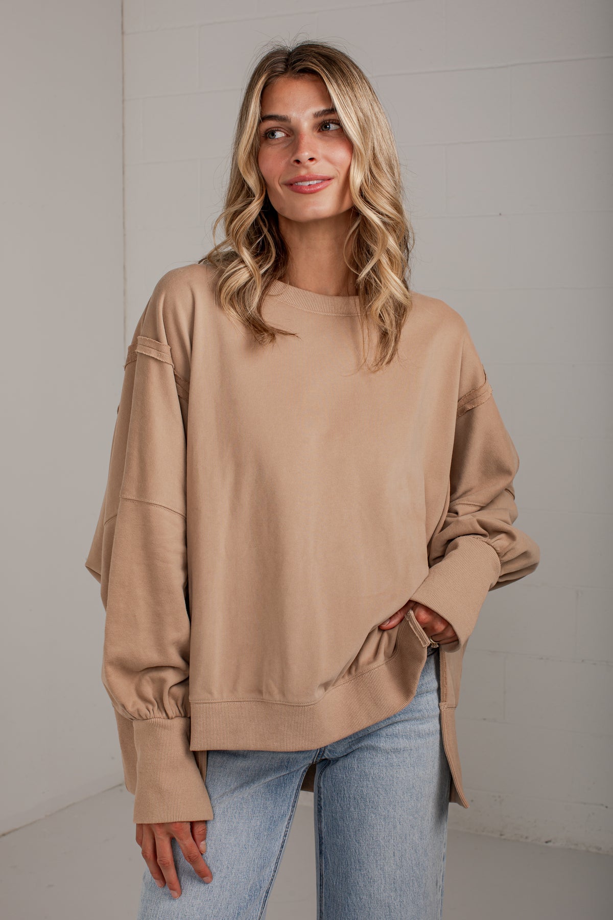A woman wearing an oversized beige crewneck sweatshirt with unique gathered sleeve details and side slits, paired with light blue jeans, standing against a light gray wall.