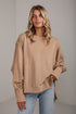 A woman wearing an oversized beige crewneck sweatshirt with unique gathered sleeve details and side slits, paired with light blue jeans, standing against a light gray wall.