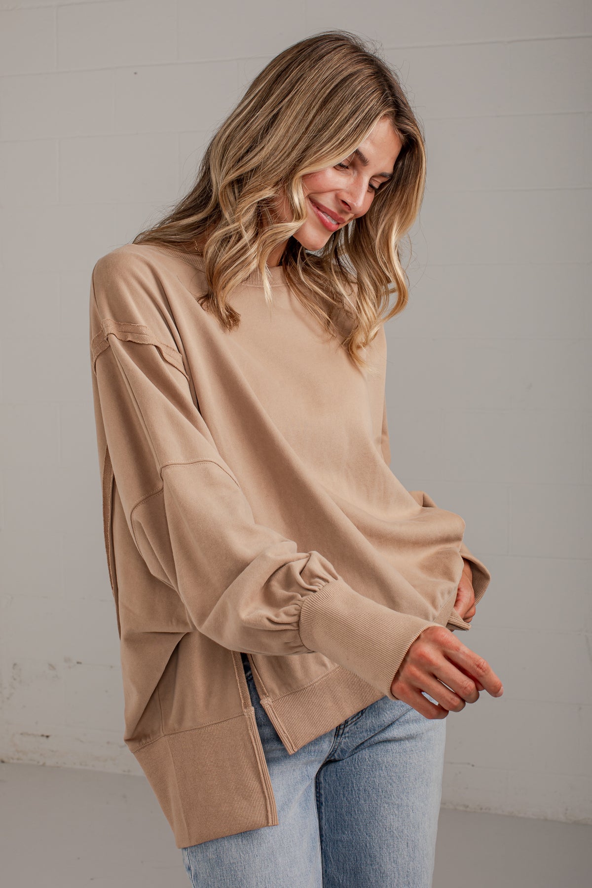 A woman wearing a beige long-sleeve knit top with gathered balloon sleeves and light blue jeans, standing against a light gray wall.