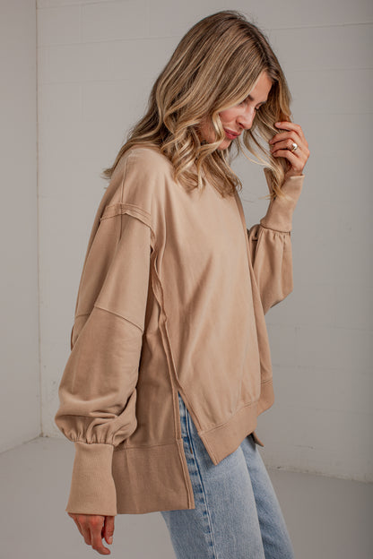 A woman wearing a loose beige long-sleeve sweatshirt with gathered balloon sleeves and light blue jeans, standing against a light gray wall.