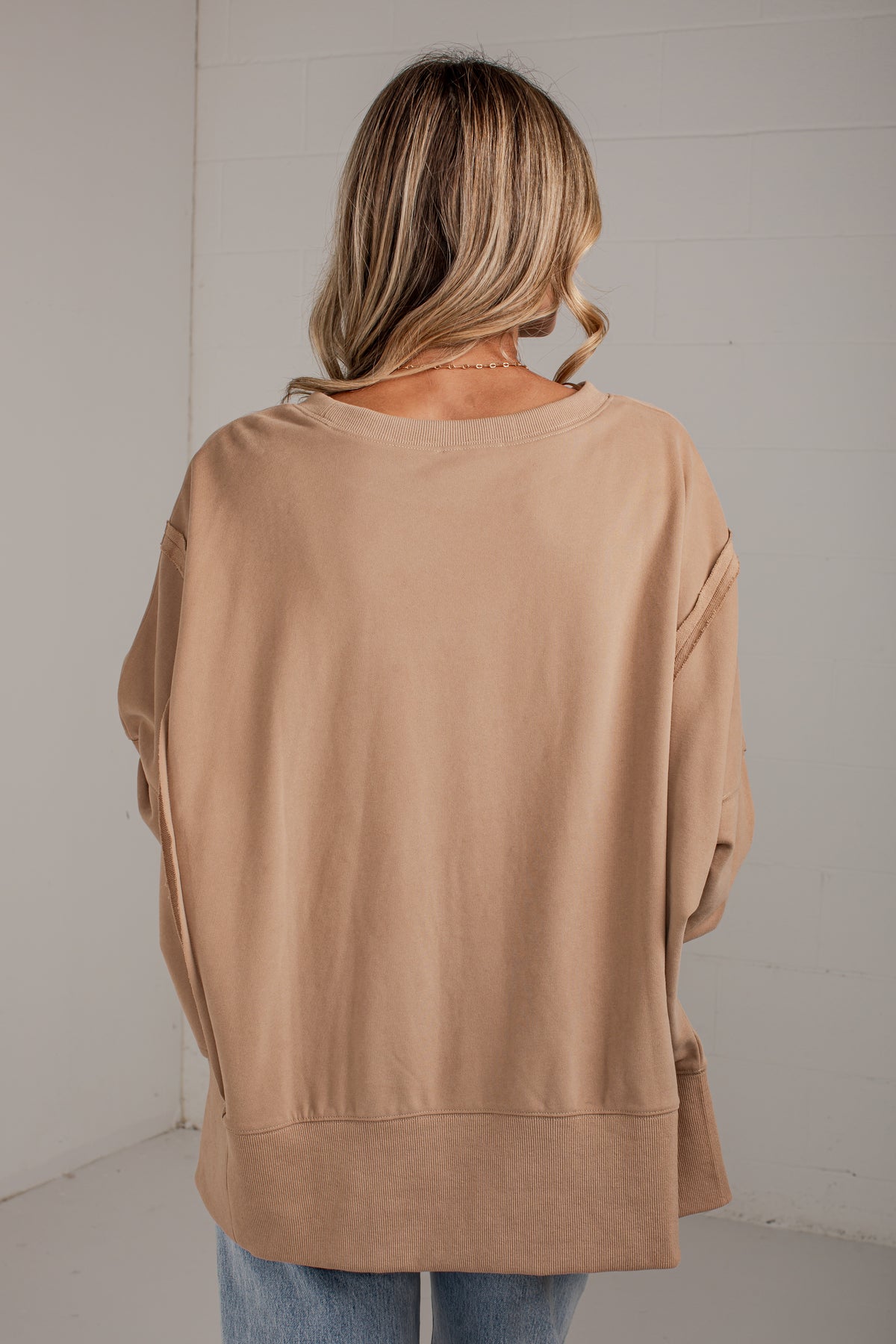 A woman viewed from behind wearing a loose, oversized tan crewneck sweatshirt and light blue jeans, standing against a white paneled wall.