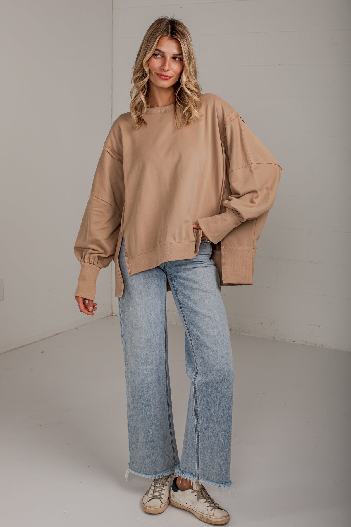 A woman modeling an oversized tan crewneck sweatshirt with balloon sleeves, paired with light blue wide-leg jeans and beige sneakers, standing against a white studio wall.