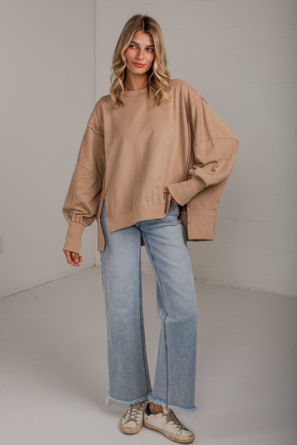 A woman modeling an oversized tan crewneck sweatshirt with balloon sleeves, paired with light blue wide-leg jeans and beige sneakers, standing against a white studio wall.
