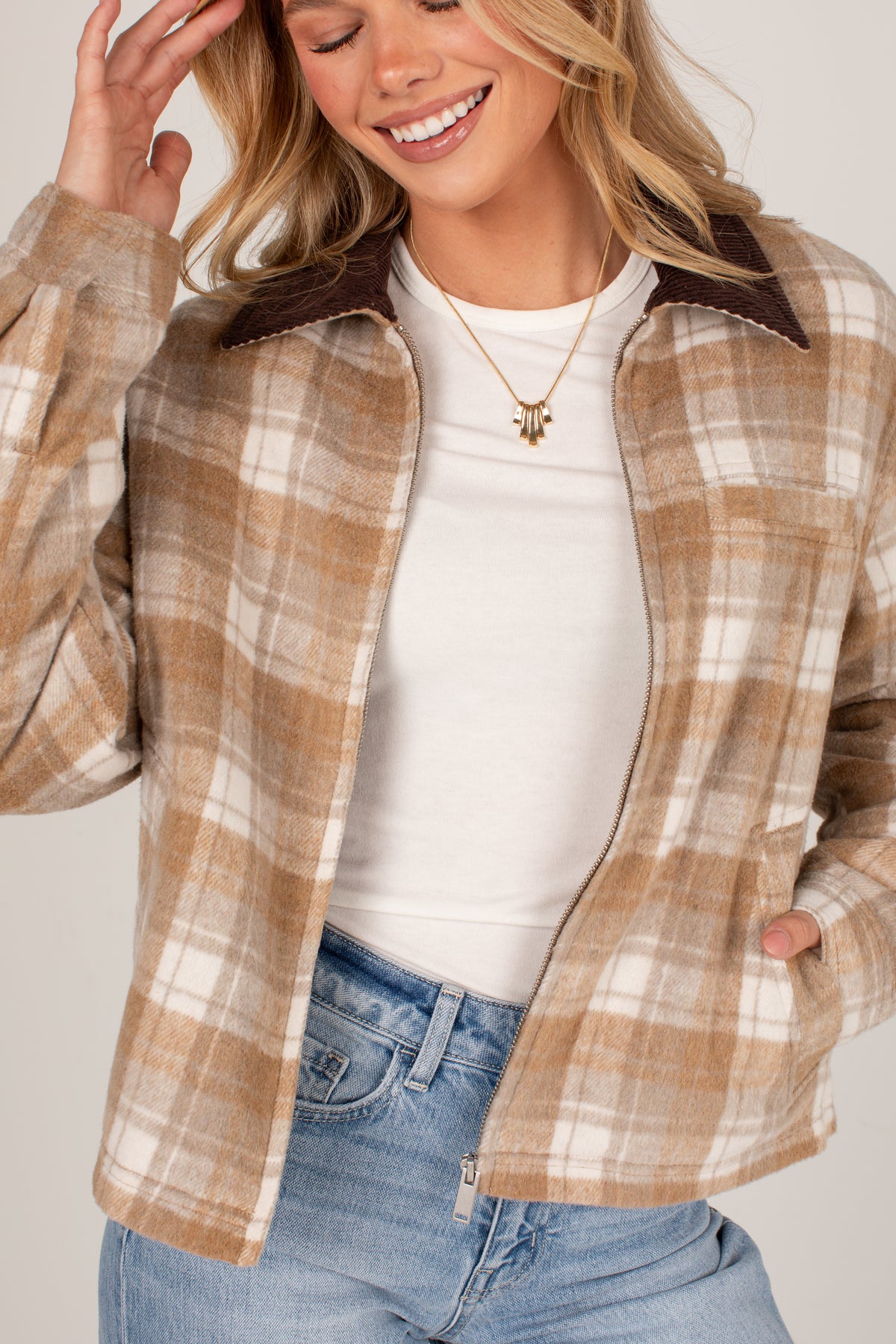 A woman wearing a tan and cream plaid button-up flannel shirt jacket with dark brown collar lining, layered over a white t-shirt and blue jeans.