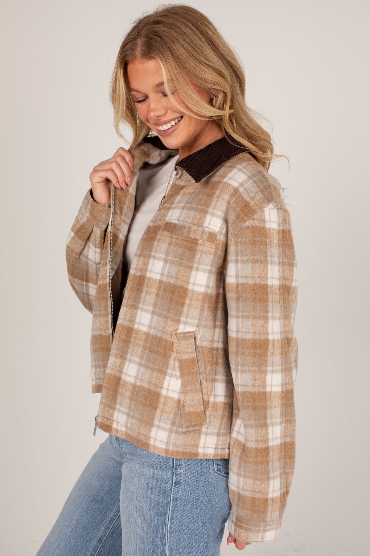A woman wearing a tan and cream plaid button-up flannel shirt jacket with a dark brown corduroy collar, paired with light blue jeans, against a plain light gray background.