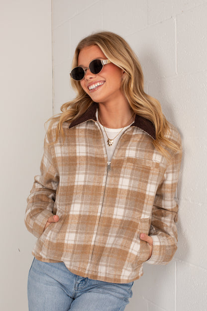A woman wearing a tan and cream plaid zip-up jacket with a dark brown collar, paired with blue jeans and sunglasses, posing against a white brick wall.
