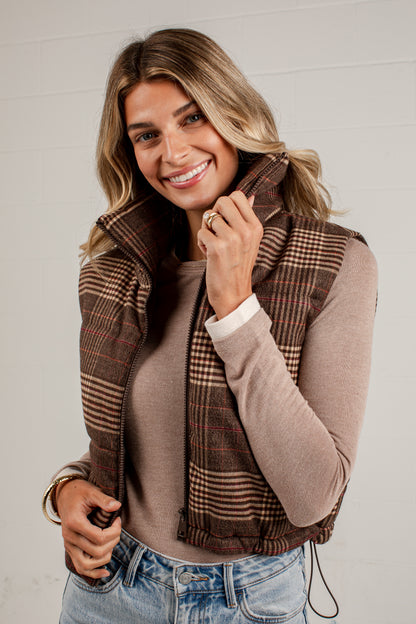 A smiling woman wearing a brown and beige plaid sleeveless puffer vest over a taupe long-sleeve top and blue jeans, standing against a light gray paneled wall.