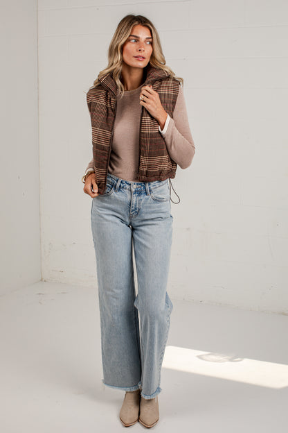 A woman modeling a brown and beige plaid sleeveless vest over a beige top, paired with light-wash wide-leg jeans and beige ankle boots, standing in a bright white studio.