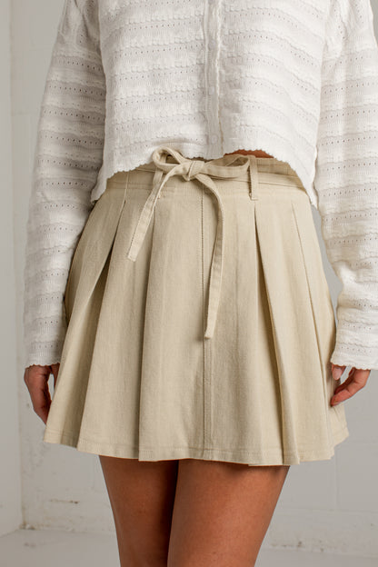 A person wearing a beige pleated mini skirt with a matching fabric tie belt at the waist, paired with a white textured knit top. The photo is cropped from the chest down against a light neutral wall.