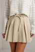 A person wearing a beige pleated mini skirt with a matching fabric tie belt at the waist, paired with a white textured knit top. The photo is cropped from the chest down against a light neutral wall.