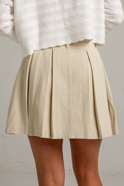 A person wearing a beige pleated mini skirt and a white textured knit top, shown from the chest down against a light neutral wall.