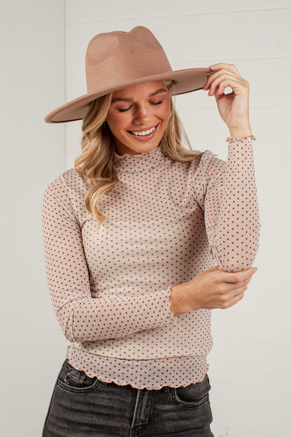 A woman wearing a beige wide-brim fedora hat and a beige long-sleeve mock neck top with a small brown polka dot pattern, paired with dark gray jeans, posing against a white brick wall.