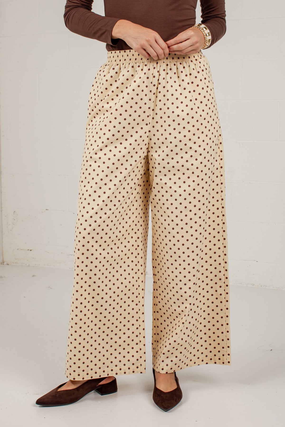 A person wearing high-waisted, wide-leg trousers with a beige base and small brown polka dots, styled with a brown long-sleeve top and brown pointed flats against a plain off-white wall.