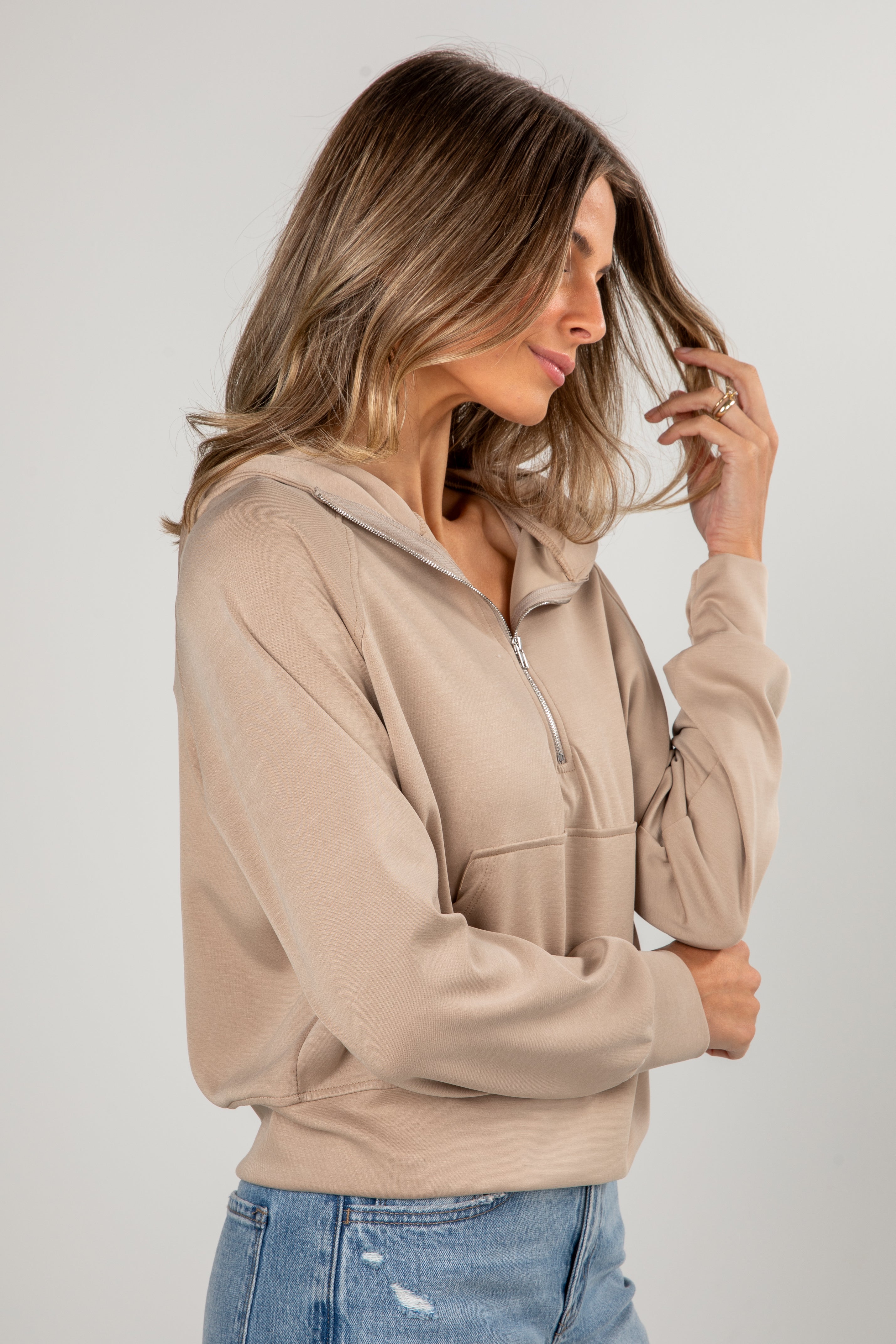 Woman wearing a beige zip-up hoodie and blue jeans against a plain background