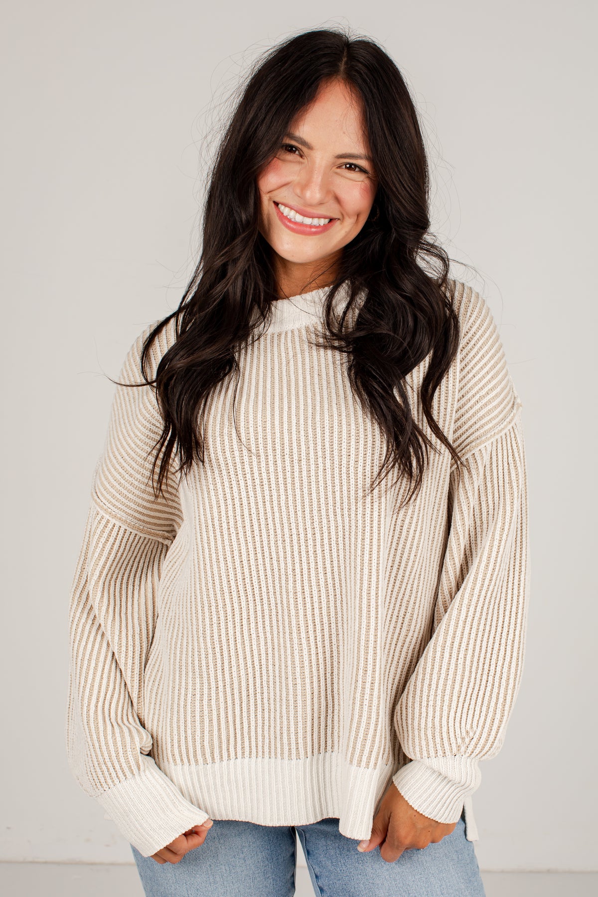 A smiling woman with long dark hair wearing a cream and beige vertically ribbed knit sweater with balloon sleeves, paired with light blue jeans, against a plain light gray background.