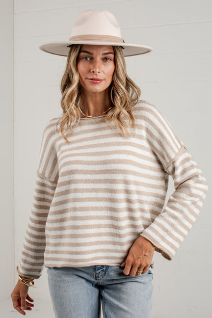A woman wearing a beige and cream striped knit sweater, a wide-brimmed cream hat, and light blue jeans, standing against a white paneled wall.
