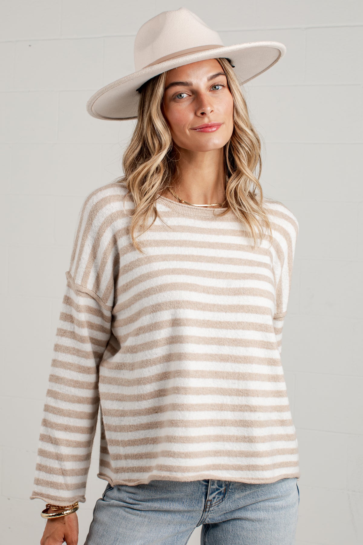A woman wearing a beige and off-white horizontally striped knit sweater, a cream wide-brim fedora hat, and light blue jeans, standing against a white paneled wall.
