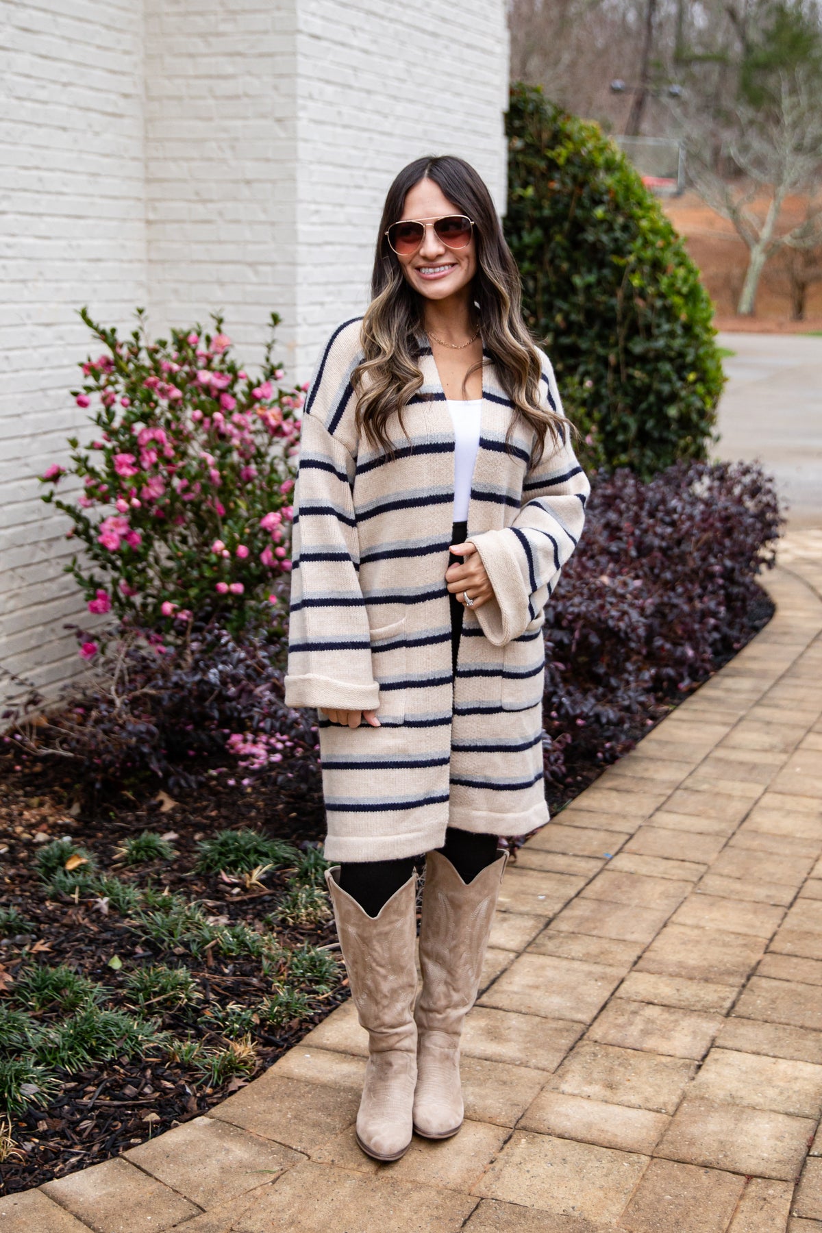 Woman wearing a striped coat and beige boots standing on a sidewalk with plants and flowers in the background.