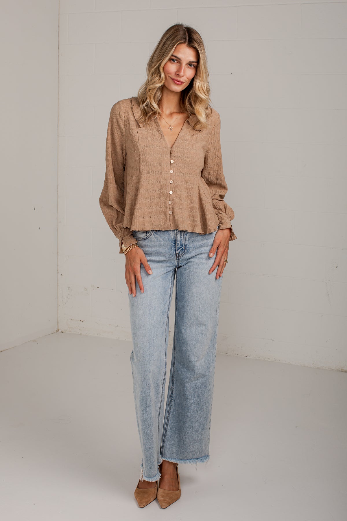 A woman modeling a light brown long-sleeve button-up blouse with a peplum hem and light-wash wide-leg jeans, standing against a plain white studio wall.