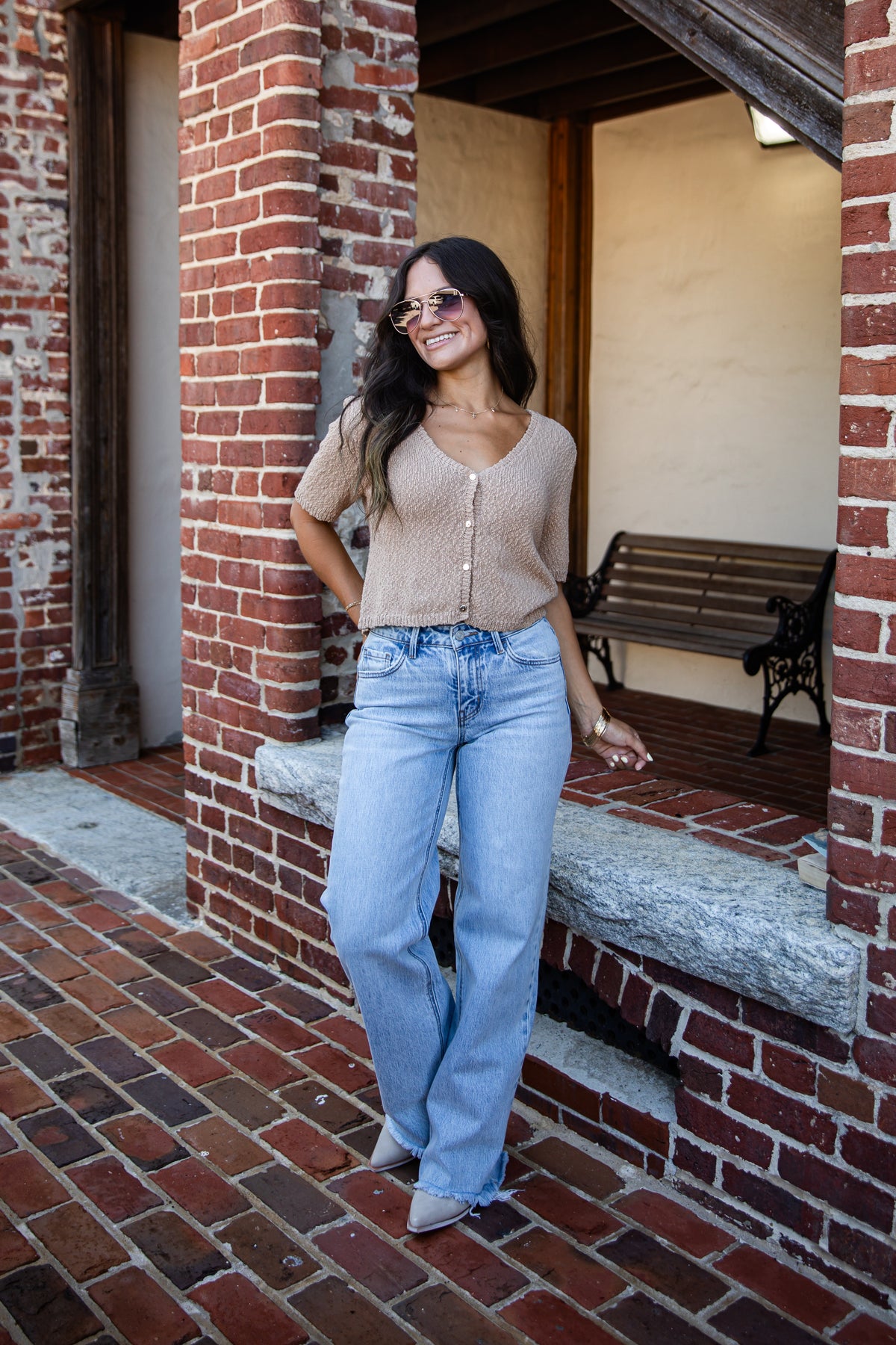 A woman wearing a beige knit V-neck top and light blue wide-leg jeans stands in a brick walkway next to a building with a wooden bench in the background.