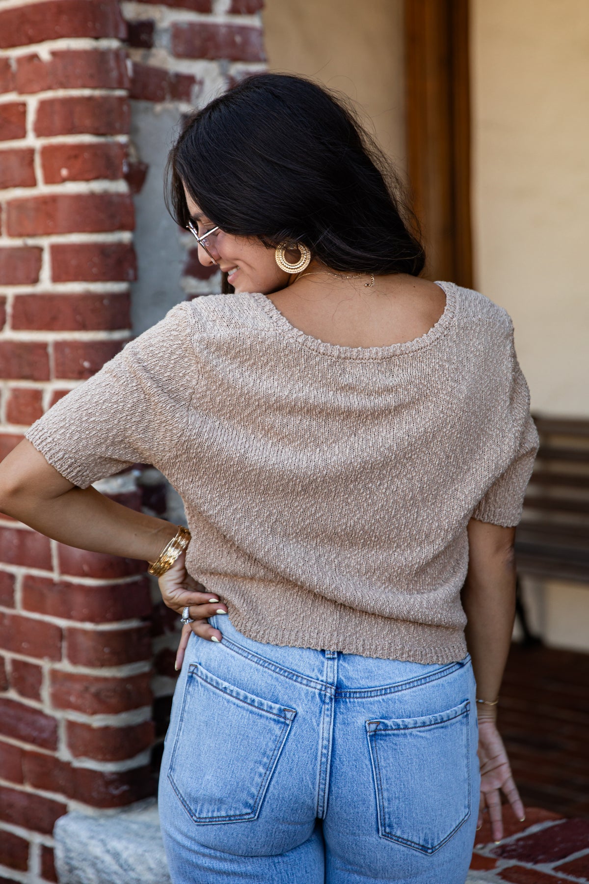A woman wearing a beige short-sleeve knit top and light blue jeans, photographed from behind in an outdoor setting with a brick wall and wooden door in the background.