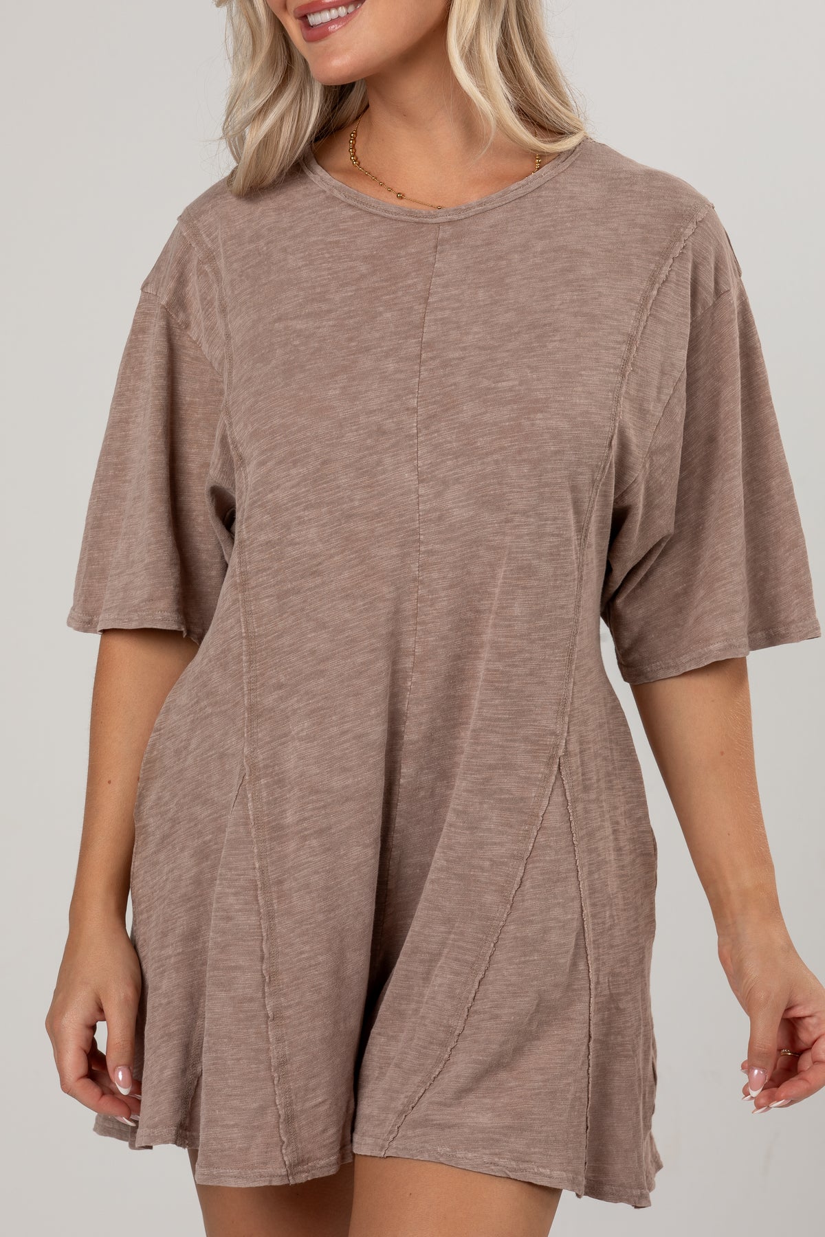 A woman wearing a loose, short-sleeve romper in a heathered taupe-brown color, shown from the waist up against a plain light gray studio background.
