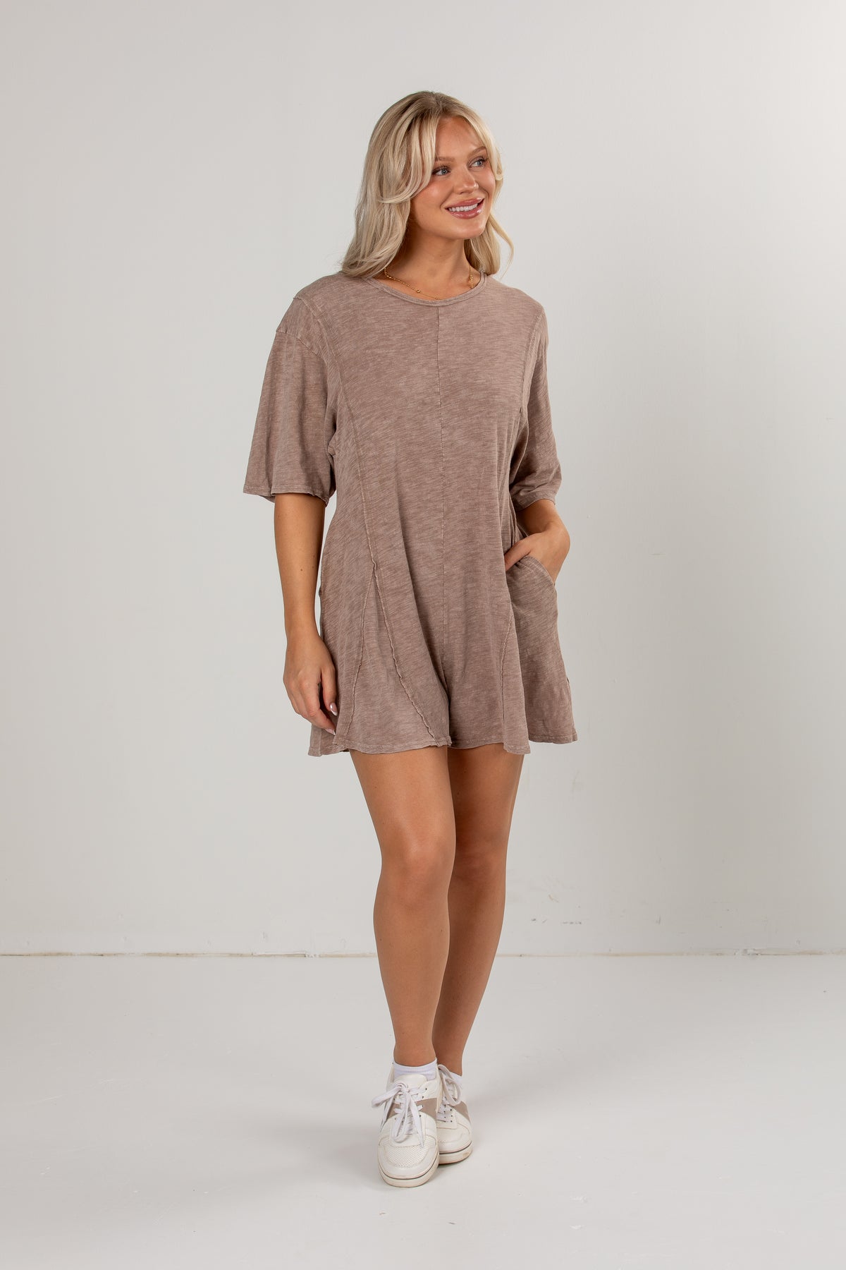 A woman modeling a short-sleeve, loose-fit mini romper in a heathered taupe-brown color, paired with white sneakers, against a plain white studio background.