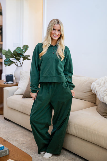 A woman stands in a living room wearing a matching dark green lounge set consisting of a long-sleeve henley-style top and wide-leg pants. The background features a beige sofa, a potted plant, and a neutral wall.