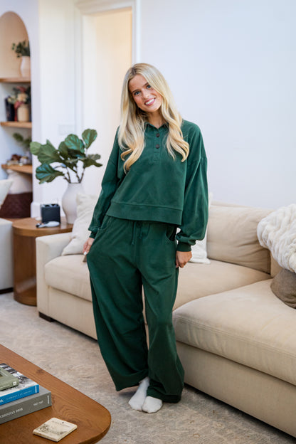 A woman stands in a living room wearing a matching dark green lounge set consisting of a long-sleeve henley sweatshirt and wide-leg sweatpants, paired with white socks.