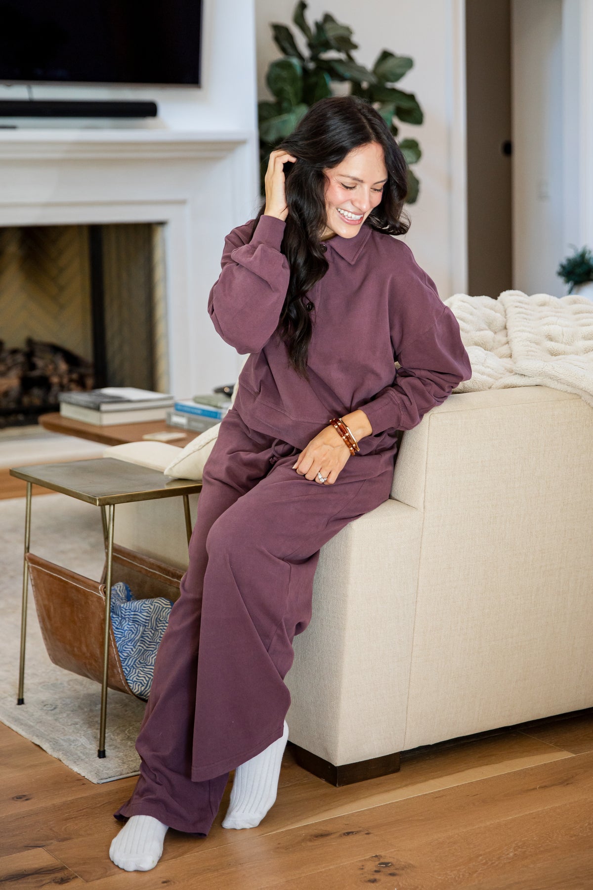 A woman wearing a matching mauve-purple lounge set consisting of a long-sleeve top and wide-leg pants, sitting on a beige sofa in a living room.